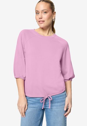 Femme portant un haut à manches longues violet clair avec un ourlet noué à l'avant, associée à un jean bleu clair, debout devant un fond uni.