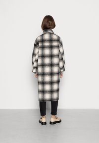 Lang, løstsiddende plaidfrakke i sort, hvid og beige. Har krave og sænkede skuldre, båret over sorte bukser og beige sko.