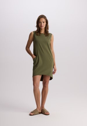 Oliwkowa sukienka bez rękawów wykonana z gładkiego materiału, z bocznymi kieszeniami i zaokrąglonym dołem, w zestawie z beżowymi sandałami na płaskiej podeszwie.