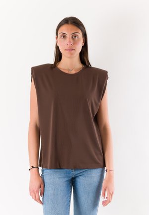 Donna con lunghi capelli scuri che indossa un top marrone senza maniche con spalle imbottite e jeans azzurri, in piedi su uno sfondo bianco neutro.