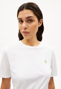 Wit katoenen t-shirt met een ronde hals, voorzien van een klein groen en goud geborduurd ontwerp van een klavertje vier op de linkerborst.