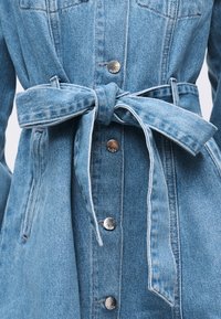 Φωτεινό γαλάζιο τζιν μπουφάν με ζώνη στη μέση που δένει και ασημένια κουμπιά με σούστες κατά μήκος του μπροστινού μέρους.