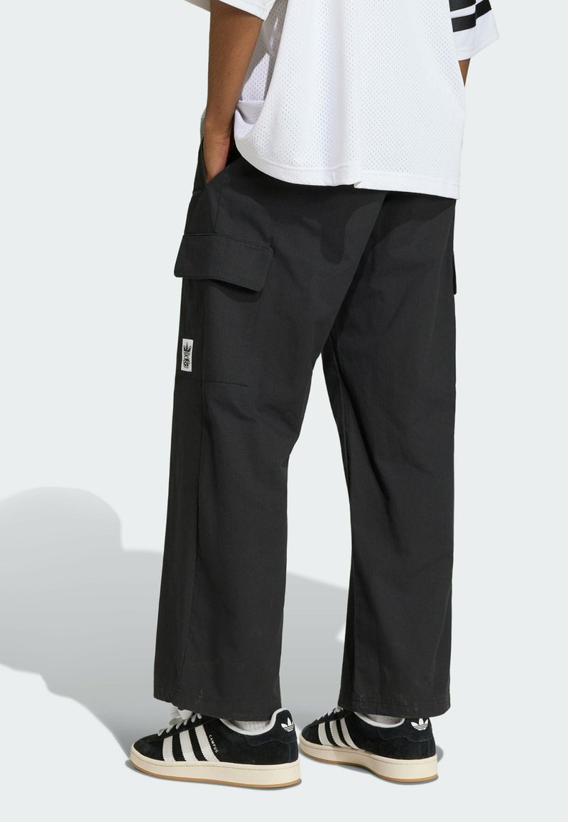 Personne portant un pantalon cargo noir ample avec des poches latérales et des baskets Adidas noires à bandes blanches, debout les mains dans les poches.