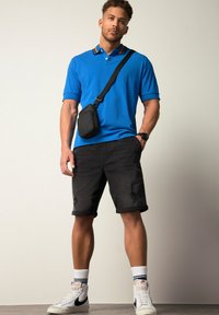 Camisa polo azul con cuello naranja y amarillo, pantalones cortos de mezclilla negros con detalles desgastados, y zapatillas deportivas blancas. Pequeño bolso cruzado negro.