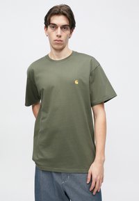 Πράσινο χακί βαμβακερό μπλουζάκι με κοντά μανίκια, στρογγυλή λαιμόκοψη και μικρό κίτρινο λογότυπο στο στήθος, με άνετη γραμμή.