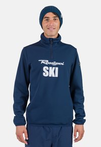 Pullover in pile blu navy con colletto a mezza zip, dotato di logo bianco "Rossignol SKI" e polsini elastici per una vestibilità aderente. Berretto di maglia blu.