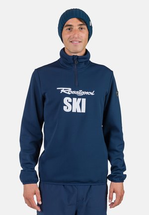 Marineblauer Fleece-Pullover mit einem halben Reißverschluss-Kragen, versehen mit einem weißen "Rossignol SKI"-Logo und elastischen Bündchen für eine eng anliegende Passform. Blauer Strickmütze.