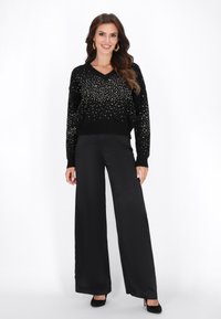 Zwarte V-hals trui versierd met zilveren embellishments, gecombineerd met hoge zwarte wijde broek en zwarte hakken.