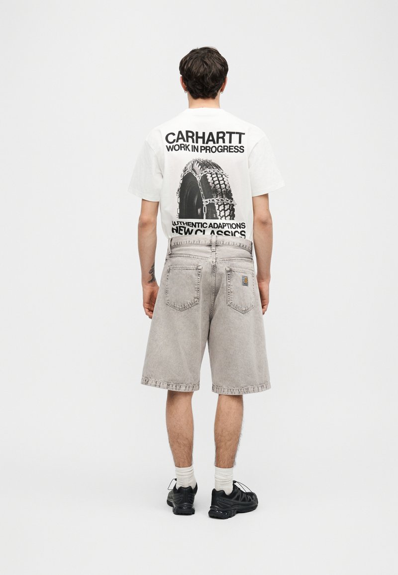 Hombre de espaldas vistiendo una camiseta gráfica blanca de Carhartt, pantalones cortos vaqueros gris claro, calcetines blancos y zapatillas negras sobre fondo blanco.