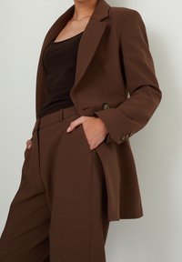 Brun kavaj i slätt tyg med strukturerade axlar, slagkrage och sidofickor; matchad med en figurnära svart linne och byxor.