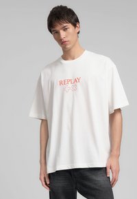 T-shirt blanc à manches courtes en tissu doux, arborant un logo "REPLAY" rouge centré avec des symboles plus et moins. Design à coupe droite.