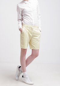 Shorts en coton jaune clair avec poches latérales, portés avec une chemise blanche à boutons ornée de petits motifs bleus et des baskets blanches.