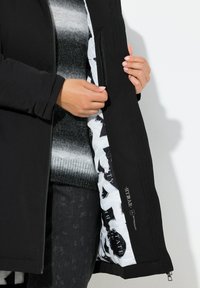 Sort jakke med tekstureret stof, der har en hvid inderforing med print. Inkluderer en lynlåslomme og et afslappet pasformdesign.