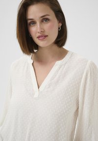 Blusa blanca ligera con puntos texturizados, un elegante escote en V y mangas largas abullonadas con sutiles puños elásticos.