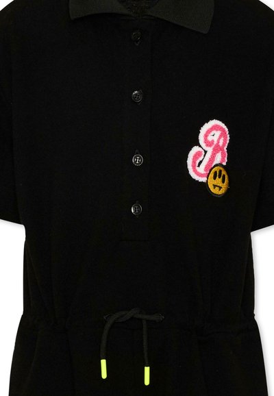 Chemise noire boutonnée avec col, lettre "R" brodée en rose et blanc, patch visage triste jaune, et cordon noir avec embouts jaune néon.