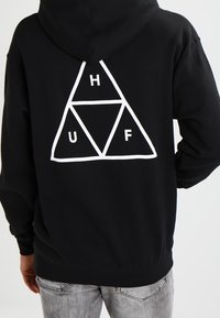 Czarna, bawełniana bluza z kapturem z dużą białą trójkątną grafiką, w której znajdują się litery H, U i F. Miękki materiał i luźny krój.