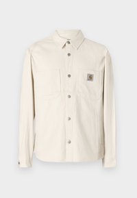 Beige színű, hosszú ujjú ingdzseki gombos elülső résszel, két mellzsebbel, ezüst színű szerkezeti elemekkel és Carhartt logo címkével a bal zsebben.
