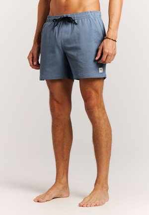 Man draagt lichtblauwe, gestructureerde zwembroek met zwarte trekkoord en een klein wit label op het linkerbeen, staat blootsvoets tegen een neutrale achtergrond.