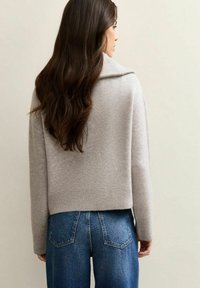 Suéter corto gris claro con cuello de chal y una textura suave, combinado con unos pantalones vaqueros ajustados de cintura alta en color azul que tienen bolsillos traseros.