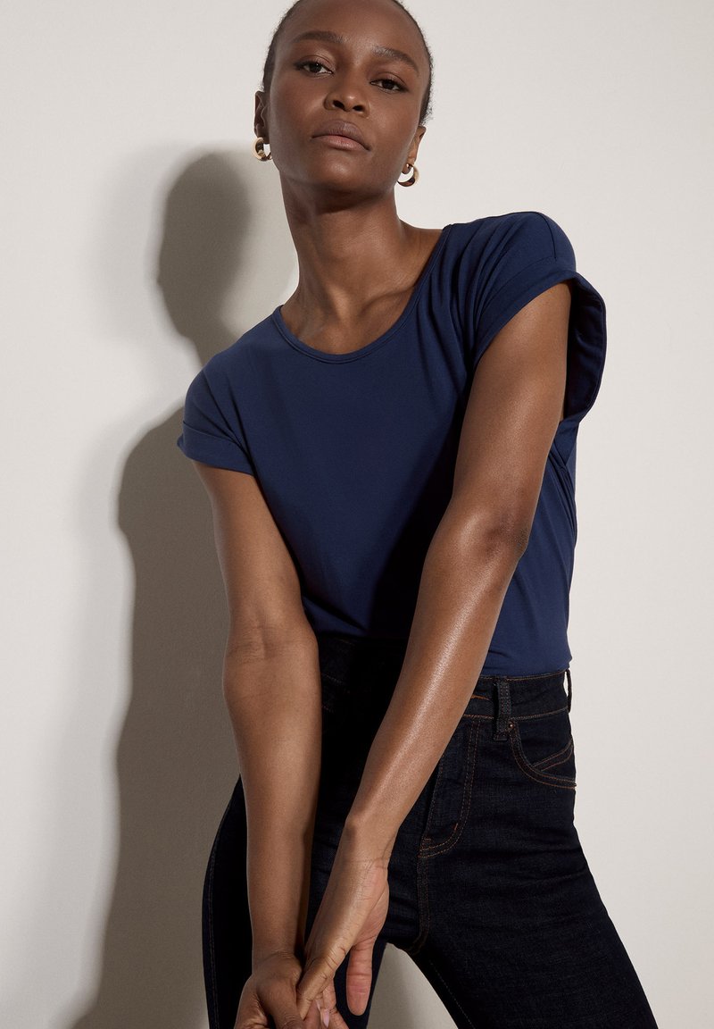 Femme aux cheveux courts portant des boucles d'oreilles créoles dorées, un haut bleu marine à manches courtes et un jean noir, debout les mains jointes devant.