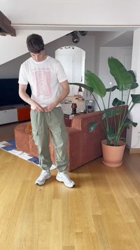 Witte graphic t-shirt met rode tekst, olijfgroene cargobroek met zakken en witte sneakers. Binnenomgeving met een plant en meubels.