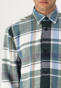 Checkered button-up shirt featuring green, blue, white, and brown colors. Textured fabric with a classic collar and black buttons.