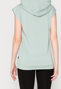 Hoodie senza maniche di colore verde menta, con vestibilità rilassata, tasche frontali e texture morbida, caratterizzato da un grande cappuccio e un orlo leggermente allungato sul retro.