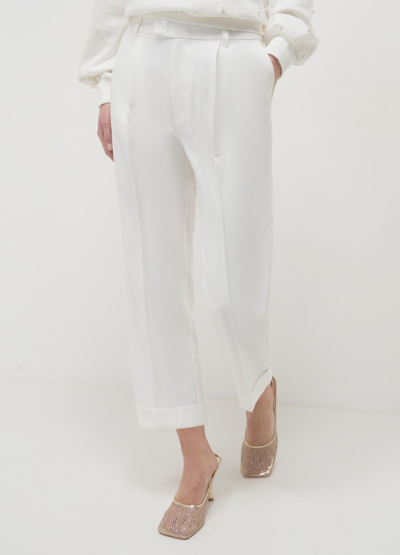Pantalon blanc sur mesure avec une texture lisse, présentant un pli marqué à l'avant, associé à des escarpins à bout ouvert métalliques.
