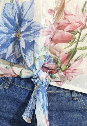Cvetlična bluzica iz prosojnega materiala z modrimi in roza cvetovi, gumbi in zavezanjem na pasu, v kombinaciji z jeans hlačami.