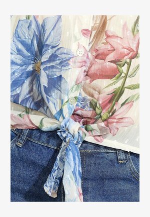 Floral blouse in sheer fabric features blue and pink flowers with button detail and a knotted tie at the waist, paired with denim jeans.