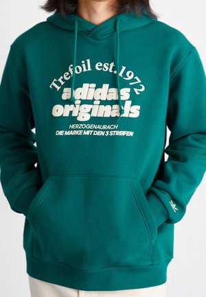 Persona que lleva una sudadera con capucha Adidas Originals verde con bolsillo delantero y texto de logo blanco, incluyendo "Trefoil est. 1972" y frase en alemán.