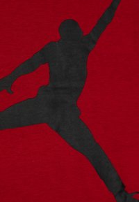 Rotes Baumwoll-T-Shirt mit einer markanten schwarzen Silhouette eines springenden Athleten, das eine glatte Textur und minimale Designelemente zeigt.