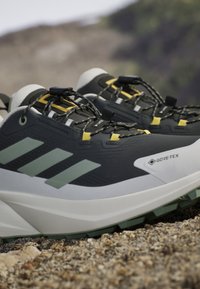 Czarne i szare buty do biegania szlakiem Adidas Gore-Tex z żółtymi akcentami na skalistym, zewnętrznym terenie.