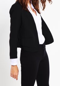 Mustat lyhyet bleiserit, joissa on muotoiltu malli, kaksi etutaskua ja sileä kangas, yhdistetty valkoiseen nappipaitaan.