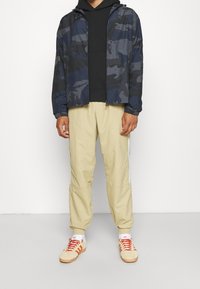 Marinblå kamouflagejacka med dragkedja och elastiska muddar, svart huva under, beige avsmalnande byxor med sidoremmar och creamfärgade sneakers med röda snören.