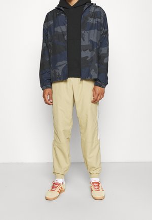 Laivastonsinisestä maastokuvioidusta hupullisesta vetoketjullisesta takista, jossa on joustavat hihansuut, musta huppari alla, beiget kujistetut housut sivuraitoineen ja vaaleat tennarit punaisilla nauhoilla.