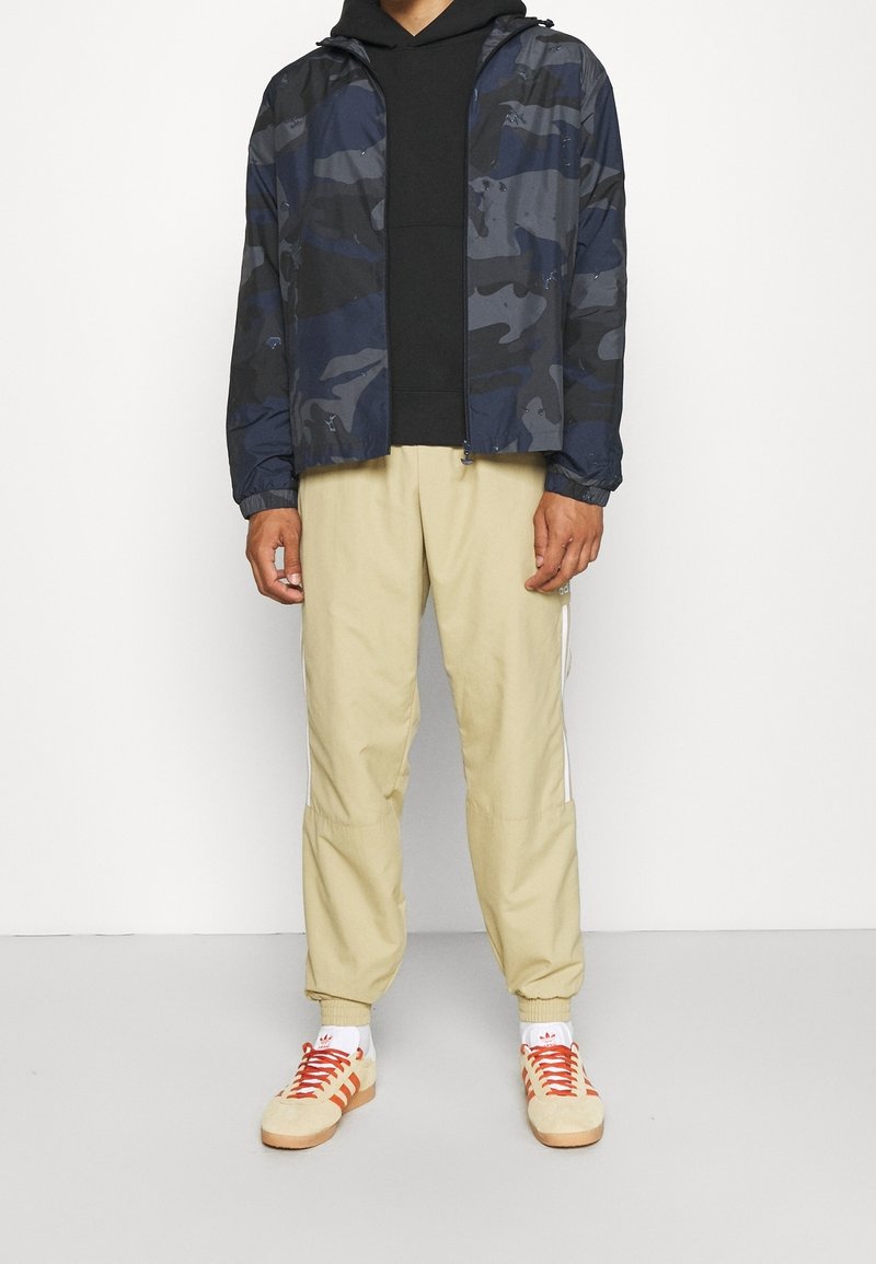 Marinblå kamouflagejacka med dragkedja och elastiska muddar, svart huva under, beige avsmalnande byxor med sidoremmar och creamfärgade sneakers med röda snören.