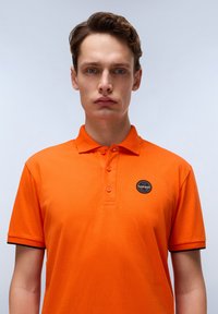 Oranžové polo triko vyrobené z bavlny, s límečkem, třemi knoflíky a kulatou nášivkou s černými akcenty na okrajích rukávů.