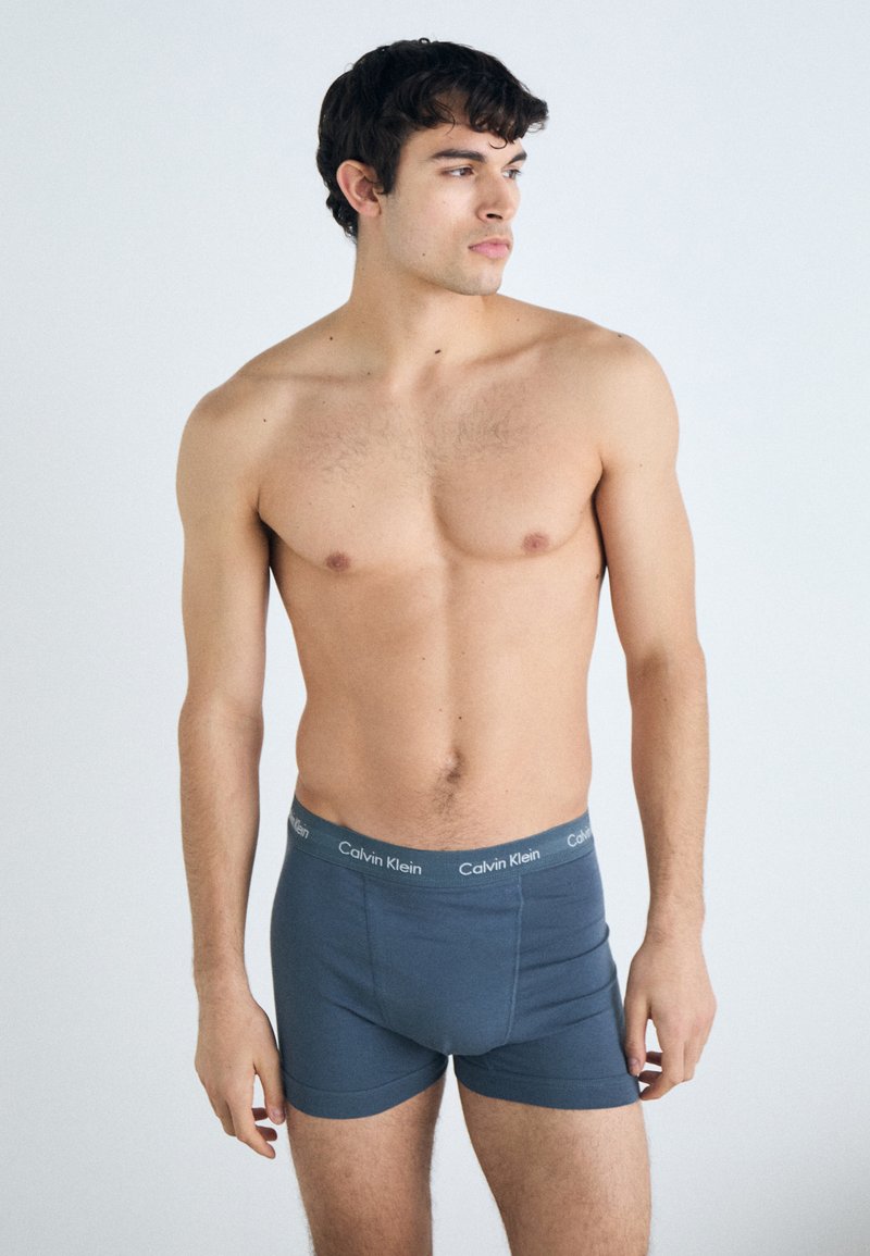 Modre bombažne bokserice z udobnim prileganjem in elastičnim pasom, na katerem je sivo napisano ime Calvin Klein. Model stoji nekoliko obrnjen v desno.