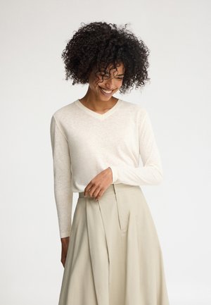 Smilende kvinde med krøllet hår iført en hvid, langærmet V-hals top, stoppet ned i et beige, højtaljet skørt, poserer mod en ensfarvet baggrund.