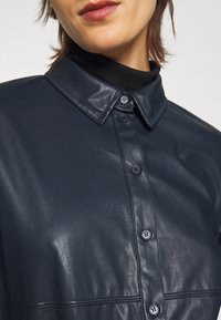 Chemise en cuir bleu marine avec une finition brillante, dotée d'un col pointu et de trois boutons sur le devant, mettant en valeur son design élégant.