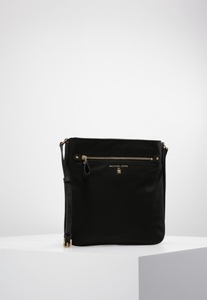 Sac bandoulière noir Michael Kors avec poche avant zippée, matériel doré et sangle d'épaule réglable sur un piédestal blanc.
