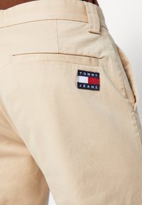Beige pants featuring a fabric patch with "TOMMY JEANS" logo, stitched with red and blue accents. Back pocket visible. Smooth texture.