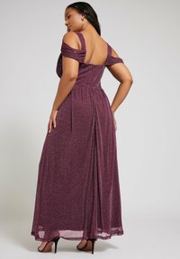 Lång, off-shoulder aftonklänning i djupt bourgogny shimmer tyg. Har en figurnära bodice, flödande kjol och bakre dragkedjestängning. Höga klackar synliga.