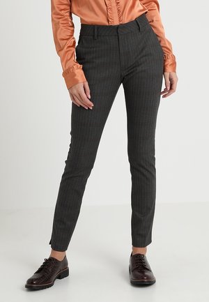 Mujer que lleva pantalones ajustados de raya diplomática gris oscuro, zapatos oxford marrones y una blusa naranja brillante de manga larga con puños fruncidos.