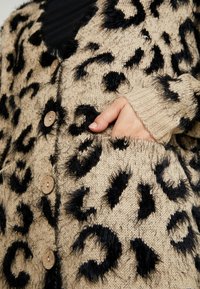 Beige gestrickte Strickjacke mit schwarzen Leopardenmustern, die flauschige Texturen und Holzknöpfe aufweist. Mit Taschen und gerippten Ärmelbündchen.