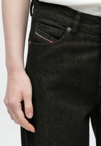 Tmavosivé denimové džínsy majú klasický dizajn s piatimi vreckami, pričom z predného vrecka vyčnieva značková štítok a sú viditeľné detaily prešívania.