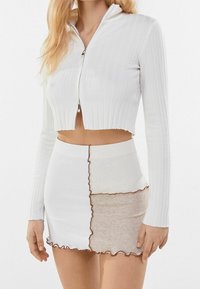 Bílý ribbed zkrácený zip-up top s dlouhými rukávy, spárovaný s béžovou a bílou mini sukní s vlnkovanými okraji a kontrastními švy.