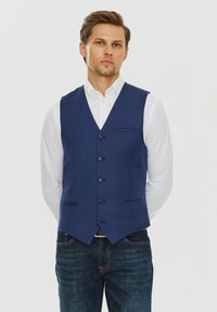 Granatowa kamizelka z fakturowanej tkaniny, z dekoltem w serek, pięcioma czarnymi guzikami oraz dwiema przednimi kieszeniami. Noszona na białej koszuli i dżinsach.