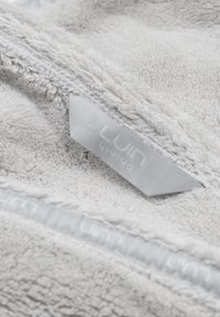 Ljusgrå handduk med en texturerad, fluffig yta. Har en vit etikett med "Luin Living" och en grå dragkedja längs en kant.